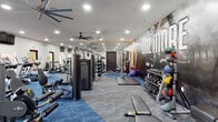 This fitness center is a comprehensive, single-floor workout facility designed to support a wide range of exercise routines. The space is anchored by a striking "ONE MORE" motivational mural that spans an entire accent wall, featuring bold athletic imagery that sets an energizing tone throughout the room. Blue carpet tile flooring defines the main workout zones, providing a vibrant visual contrast against the light gray walls and white ceiling.

The strength training area is extensively outfitted with TRUE-brand cable and selectorized machines, multiple dumbbell racks with a full range of free weights, medicine balls, barbells, and power racks with benches. Full-length mirrors line one wall, enhancing the sense of depth and allowing users to monitor their form during lifts. Padded exercise mats are available for floor-based work.

The cardio section features a row of treadmills, ellipticals, stair climbers, and stationary bikes, all positioned near windows with views to the outdoors. Mounted flat-screen televisions are positioned above the cardio equipment for entertainment. A glass-panel door provides direct outdoor access from this area.

Oversized ceiling fans are distributed throughout the space, ensuring consistent airflow during workouts. Recessed lighting provides bright, even illumination across the entire facility, creating a clean and professional gym environment.
