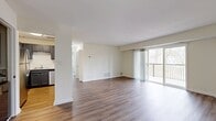 This 940-square-foot, 2-bedroom, 1-bathroom apartment is located at Commons at Windsor Gardens in Norwood, MA. The open-concept living room features light hardwood-style flooring, neutral walls, and large sliding glass doors that open to a private balcony with wooded views, filling the space with abundant natural light. The kitchen is well-appointed with gray cabinetry, stainless steel appliances including a refrigerator, electric range, and built-in microwave, complemented by a white tile backsplash and light countertops. A convenient pass-through counter connects the kitchen to the living area. In-unit stacked laundry is also visible off the kitchen. The primary bedroom spans 190 square feet and features large windows with tree-lined views and hardwood-style flooring. The second bedroom is similarly bright with a window overlooking greenery. Both bedrooms include closets with built-in shelving and hanging rods. The full bathroom is finished in white with a tub/shower combo, white vanity, and coordinating hardwood-style flooring. Throughout the unit, clean lines and a neutral palette create a bright, cohesive living environment.