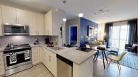 This well-appointed 2-bedroom, 2-bathroom residence is designed with a contemporary open-concept floor plan that seamlessly connects the kitchen, dining, and living areas. The modern kitchen is outfitted with crisp white cabinetry, a sleek quartz countertop peninsula that doubles as a breakfast bar, a stainless steel undermount sink, and stainless steel appliances including a dishwasher and range. A stylish tile backsplash and pendant lighting add refined detail overhead, while a track lighting system provides flexible illumination throughout the space. Light wood-style flooring runs throughout the main living area, contributing to a clean and airy aesthetic. The living space makes a bold design statement with a deep navy blue accent wall that anchors the room, complemented by a neutral sofa adorned with yellow and blue throw pillows. A framed artwork piece and a sculptural floor lamp further enhance the curated ambiance. A glimpse into the second bedroom reveals a continuation of the vibrant color palette. The overall design balances modern finishes with bold, expressive touches, creating a space that feels both stylish and functional.