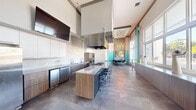 Located at 20080 West Dixie Highway in Miami, FL, this single-floor community space is a thoughtfully designed culinary and social hub. The open-concept layout is anchored by a generously sized central kitchen island topped with a sleek countertop, surrounded by modern chrome bar stools and equipped with a professional-grade gas cooktop and overhead stainless steel range hood. The kitchen wall features full-size stainless steel appliances, including a large refrigerator, double wall ovens, an undercounter beverage cooler, and dual sinks — all complemented by a combination of white upper cabinetry and warm wood-toned lower cabinets. A wall-mounted flat-screen TV adds a multimedia element to the kitchen zone. The space transitions seamlessly into a lounge and dining area, where a striking pendant chandelier with Edison-style bulbs hangs above a grouping of modern chairs and a meeting table. An accent wall clad in gradient teal mosaic tile adds vibrant color and artistic character to this corner. Large floor-to-ceiling windows along one wall flood the interior with abundant natural light and offer views of the outdoor surroundings. A built-in counter beneath the windows provides additional workspace or casual seating. Curated artwork, stone-look tile flooring, and soaring ceilings complete the polished, contemporary ambiance of this versatile gathering space.