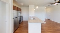 This single-floor residence offers a thoughtfully designed layout that balances open communal spaces with comfortable private retreats. The kitchen serves as a central highlight, featuring rich dark wood cabinetry, granite-look countertops, and a generous island with a built-in double sink and pendant lighting overhead. Stainless steel appliances, including a refrigerator, microwave, and range, complete the well-equipped cooking space. Warm hardwood-style flooring flows throughout the kitchen and living areas, lending a cohesive and inviting feel to the open-concept layout. The bedrooms are finished with plush carpeting and benefit from natural light through slatted blinds. One bedroom includes a ceiling fan and a view to an outdoor balcony, while another features a spacious walk-in closet with built-in shelving for ample storage. The bathroom is appointed with classic white subway tile surrounding a full soaking tub and shower combination, accented by a contrasting tile band. Hardwood-style flooring continues into the bathroom, complementing the clean, white walls and brushed nickel fixtures. Throughout the space, recessed lighting and neutral tones create a bright, modern ambiance suited to a variety of lifestyles.
