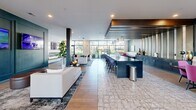 Located at 11005 Octave Dr in Zionsville, Indiana, this expansive single-floor communal space offers a thoughtfully designed open-concept layout that seamlessly blends multiple functional zones. The interior is defined by a bold, contemporary aesthetic, featuring a striking color palette of deep teal accent walls, crisp whites, and warm wood tones throughout.

The living area serves as the heart of the space, anchored by a sleek wall-mounted fireplace beneath a large flat-screen display, and furnished with plush white sofas, tufted leather benches, and layered area rugs. Adjacent to this is a social lounge zone featuring a long bar counter with a dark navy finish, complemented by upholstered bar stools and elegant pendant lighting suspended from a dramatic wood-slat ceiling feature above.

A decorative geometric-patterned tile floor transitions into warm hardwood, visually distinguishing different seating and activity areas. Natural light floods the space through expansive floor-to-ceiling windows along one wall, offering views of the surrounding outdoor landscape. A distinctive gold-framed glass partition adds an architectural focal point, while curated artwork and sculptural décor accessories enhance the upscale ambiance throughout. Recessed lighting ensures the entire space is evenly and warmly illuminated.