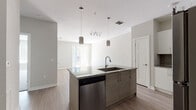 This well-appointed single-floor unit showcases a contemporary design with a neutral color palette, light wood-look flooring, and an open-concept layout that promotes a seamless flow between spaces. The kitchen serves as the centerpiece of the home, featuring a large central island topped with granite countertops and equipped with an undermount sink and pendant lighting. White upper cabinetry with sleek hardware contrasts with the darker-toned island cabinetry, while a gray subway tile backsplash adds texture and depth. A full suite of stainless steel appliances — including a refrigerator, electric range, over-the-range microwave, and dishwasher — completes the functional and stylish kitchen. The open living area extends naturally from the kitchen, bathed in natural light from large windows and sliding glass door access. A ceiling fan adds comfort to the living space, which benefits from a clean, airy atmosphere. The bathroom features a full tub-shower combination with tile surround, a granite-topped vanity with white cabinetry, and wood-look flooring that ties in with the rest of the unit. Throughout the space, textured ceilings, crisp white trim, and consistent finishes contribute to a cohesive and modern aesthetic.