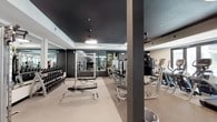 This fitness center is thoughtfully designed to accommodate a wide range of workout styles across a single, expansive floor. The space is defined by its striking dark-painted ceilings contrasted against white crown molding and light-toned walls, creating a sleek, modern aesthetic throughout. Durable speckled flooring covers the main workout areas, offering both practicality and visual consistency.

The strength training zone is well-equipped with a full dumbbell rack, cable machines, a multi-functional power rack, and adjustable benches. Large wall-mounted mirrors line multiple sections, enhancing the sense of depth and allowing users to monitor their form. Slatted wall panels provide organized storage for accessories such as resistance bands, medicine balls, and other training tools.

The cardio area features a row of treadmills and elliptical machines positioned near windows, offering views to the outdoors. Ceiling fans and round flush-mount light fixtures ensure comfortable, well-lit conditions throughout.

A dedicated functional training room offers open floor space with a spin bike, stability balls, parallette bars, and plyo boxes, with French doors that open to an outdoor courtyard. A reception-style shelving unit near the entrance adds organizational functionality to the overall space.