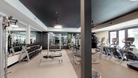 This fitness center is thoughtfully designed to accommodate a wide range of workout styles across a single, expansive floor. The space is defined by its striking dark-painted ceilings contrasted against white crown molding and light-toned walls, creating a sleek, modern aesthetic throughout. Durable speckled flooring covers the main workout areas, offering both practicality and visual consistency.

The strength training zone is well-equipped with a full dumbbell rack, cable machines, a multi-functional power rack, and adjustable benches. Large wall-mounted mirrors line multiple sections, enhancing the sense of depth and allowing users to monitor their form. Slatted wall panels provide organized storage for accessories such as resistance bands, medicine balls, and other training tools.

The cardio area features a row of treadmills and elliptical machines positioned near windows, offering views to the outdoors. Ceiling fans and round flush-mount light fixtures ensure comfortable, well-lit conditions throughout.

A dedicated functional training room offers open floor space with a spin bike, stability balls, parallette bars, and plyo boxes, with French doors that open to an outdoor courtyard. A reception-style shelving unit near the entrance adds organizational functionality to the overall space.