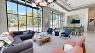 Located at 335 E Duval Street in the heart of downtown Jacksonville, Florida, this contemporary single-floor communal space offers a thoughtfully designed environment that balances open social gathering with intimate seating arrangements. Soaring ceilings and expansive floor-to-ceiling black-framed windows dominate the exterior-facing walls, flooding the interior with abundant natural light and framing lush views of the surrounding mature oak trees and streetscape.

The open-plan layout is organized into distinct functional zones. A generous lounge area features a large sectional sofa, accent chairs in muted tones of pink and blue, and a solid wood coffee table, all anchored by a soft area rug. Nearby, multiple dining and café-style tables with upholstered green and wood chairs provide versatile options for working or gathering. A standout feature is the built-in bar counter finished in sage green tile, accompanied by bar stools and a wall-mounted television framed by a decorative mosaic tile surround — an ideal setting for social events or casual meetings.

Curated contemporary artwork punctuates the neutral walls, complementing the polished concrete-style flooring and textured ceiling elements, including sculptural pendant lighting and an architecturally expressive ceiling fixture. The overall ambiance is modern, airy, and inviting, with a refined aesthetic that suits both professional and leisure use.