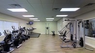 This single-floor fitness center is designed to provide a comprehensive workout experience in a clean, functional environment. The space is outfitted with a variety of cardiovascular machines, including multiple treadmills and an elliptical trainer, arranged along one side of the room for easy access and comfortable spacing between equipment. On the opposite side, a multi-station strength training machine offers versatile resistance-based workouts, complemented by a vertical knee raise and dip station for core and upper-body exercises. A flat-screen television is mounted on the wall, providing entertainment during cardio sessions. The flooring consists of durable, wood-look planks that are easy to maintain and lend a clean, modern aesthetic to the space. Overhead, a drop ceiling with large recessed panel lights ensures the room is evenly and brightly illuminated throughout. The walls are painted in a soft, neutral tone that contributes to a calm and focused workout atmosphere. The layout is thoughtfully organized to maximize usable floor space, allowing for fluid movement between stations. A private door at the rear of the room provides additional access or utility. Overall, the fitness center is well-suited for a full-body workout routine in a comfortable, well-maintained setting.