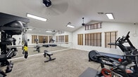 This dedicated fitness space is thoughtfully designed to support a wide range of workout styles within a single, open-plan room. The vaulted ceiling lends a sense of openness and airiness, while two ceiling fans ensure consistent air circulation throughout the space. Flush-mounted fluorescent light fixtures provide bright, even illumination across the entire room.

A standout architectural feature is the full-length wall of mirrors paired with a mounted ballet barre, which visually expands the room and makes it equally suited for strength training, stretching, or barre-style workouts. Opposite the mirror wall, a row of windows fitted with wood-toned blinds — including clerestory-level windows near the roofline — allows for natural light while maintaining privacy and light control.

The carpeted floor provides a comfortable underfoot surface throughout the space. Cardio equipment includes a treadmill, a Peloton spin bike, and an elliptical machine. A dedicated free-weight area features a tiered dumbbell rack, kettlebells, medicine balls, and resistance bands stored on a multi-level rack. A flat-screen television is mounted on the wall, and a weight bench adds versatility for strength training. The space also includes a door leading to an adjacent room, rounding out this fully functional, private fitness facility.