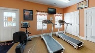 Located at Pond's Edge in Delmar, Maryland, this fitness center offers a well-equipped, single-floor space designed to support a variety of workout routines. The room is finished with warm peach-toned walls and carpeted flooring, with protective rubber mats placed beneath the equipment, creating a practical yet inviting environment.

The cardio section is well-appointed with multiple treadmills, elliptical trainers, a stair climber, and both upright and spin bikes, providing a full range of aerobic options. A multi-station strength training machine anchors the strength-training area, complemented by additional resistance equipment to support full-body workouts. Wall-mounted mirrors along one side of the room allow users to monitor their form during exercise.

Natural light filters in through several windows fitted with decorative valances, and the space is further illuminated by overhead fluorescent lighting. A ceiling fan aids in air circulation and comfort during workouts. Mounted flat-screen televisions are positioned throughout the room for entertainment while exercising. A water cooler station is conveniently available for hydration. The facility is accessible through both a standard entry door and a set of double doors, with clearly marked emergency exits ensuring safety throughout the space.