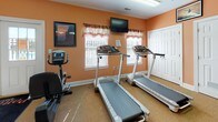 Located at Pond's Edge in Delmar, Maryland, this fitness center offers a well-equipped, single-floor space designed to support a variety of workout routines. The room is finished with warm peach-toned walls and carpeted flooring, with protective rubber mats placed beneath the equipment, creating a practical yet inviting environment.

The cardio section is well-appointed with multiple treadmills, elliptical trainers, a stair climber, and both upright and spin bikes, providing a full range of aerobic options. A multi-station strength training machine anchors the strength-training area, complemented by additional resistance equipment to support full-body workouts. Wall-mounted mirrors along one side of the room allow users to monitor their form during exercise.

Natural light filters in through several windows fitted with decorative valances, and the space is further illuminated by overhead fluorescent lighting. A ceiling fan aids in air circulation and comfort during workouts. Mounted flat-screen televisions are positioned throughout the room for entertainment while exercising. A water cooler station is conveniently available for hydration. The facility is accessible through both a standard entry door and a set of double doors, with clearly marked emergency exits ensuring safety throughout the space.