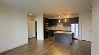 This single-floor space offers a thoughtfully designed open-concept layout that seamlessly blends the kitchen and living areas into one cohesive and functional environment. The kitchen is a standout feature, outfitted with rich dark cabinetry that provides ample storage and a bold visual contrast against the light granite countertops. A mosaic tile backsplash adds texture and dimension to the space, while stainless steel appliances — including a refrigerator, electric range, and microwave — deliver a sleek, modern finish. A center island serves as both a prep station and casual dining spot, illuminated overhead by elegant pendant lighting. Track lighting runs along the ceiling, providing flexible and targeted illumination throughout the kitchen. The flooring throughout the space is a warm-toned hardwood-style material that adds continuity and a welcoming feel underfoot. The open layout allows natural sightlines from the kitchen into the adjoining living area, enhancing the sense of spaciousness. A hallway visible from the main living space leads to additional rooms, and clean white walls paired with recessed lighting contribute to a bright, modern ambiance throughout this well-appointed single-level home.