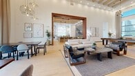 Located at 1161 E O'Neil Dr in Casa Grande, Arizona, this single-story space offers a generously sized, open-concept layout defined by striking architectural details and a cohesive contemporary aesthetic. The living area commands attention with its dramatically high ceilings accented by exposed wooden beams and recessed lighting, complemented by an array of designer pendant fixtures that provide warm, layered illumination throughout.

Light hardwood flooring in a chevron pattern anchors the space, flowing seamlessly across distinct functional zones. Floor-to-ceiling windows along one wall flood the interior with abundant natural light while framing views of the surrounding Arizona landscape, including palm trees and the desert sky. Sheer linen drapery softens the window walls without obstructing the views.

A central lounge area features a neutral sofa arrangement, leather accent chairs, and a substantial wooden console table, anchored by a fireplace with built-in shelving. An adjacent seating nook with warm cognac-toned sofas and layered textiles creates an inviting retreat. A wood-clad structural column adds warmth and visual interest, while recessed booth-style seating and counter-height workspaces contribute additional functional areas. Curated wall art and indoor greenery complete the refined, welcoming ambiance.