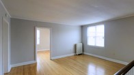 This single-floor space presents a straightforward and functional layout, characterized by warm hardwood flooring that runs throughout and lends a sense of continuity between rooms. The main living area is finished with soft gray walls and crisp white trim and crown molding, creating a clean, classic aesthetic. A traditional cast-iron radiator stands along the wall, indicative of a well-established heating system. Natural light enters through a double-hung window, brightening the space and highlighting the reflective quality of the hardwood floors. An open doorway connects to an adjacent room, which also features hardwood flooring and its own window, allowing light to flow through the space and reinforcing the open, airy feel. The white ceiling is smooth and well-maintained, complementing the neutral color palette throughout. The overall ambiance is calm and understated, offering a blank canvas that is practical and easy to furnish. The layout promotes a natural flow between spaces, making it well-suited for a variety of uses. The combination of classic architectural details, including the molding and radiator, with the timeless hardwood floors gives this space a grounded, enduring character.