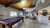 Located at 5400 N Nevada Ave in Colorado Springs, CO, this single-floor community clubhouse offers a well-appointed gathering space designed for both relaxation and productivity. The centerpiece of the space is the expansive kitchen and living room area, where soaring vaulted ceilings clad in warm wood planking and bold exposed beams create a striking architectural statement. The living area features a stacked-stone fireplace topped with a wall-mounted television, elegant chandelier lighting, and a comfortable seating arrangement ideal for socializing. A full-size billiards table anchors one side of the room, adding a recreational focal point. Multiple windows throughout invite natural light and provide views of the surrounding landscape. The kitchen is fully equipped with wood cabinetry, black appliances including a range, microwave, and refrigerator, along with ample counter space and a pass-through opening to the main living area. Adjacent to the kitchen, a casual dining zone features high-top tables and a coffee station with warm wood accent walls. The dedicated office features a row of computer workstations set along a continuous red-lacquered desk surface, complemented by a long display shelf showcasing decorative accessories. Light wood-tone flooring flows consistently throughout, unifying the entire space.