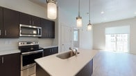 This well-appointed single-floor residence showcases a thoughtfully designed open-concept layout that seamlessly connects the kitchen and living areas. The kitchen is a standout feature, outfitted with dark espresso cabinetry extending to the ceiling for ample storage, a full suite of stainless steel appliances including a refrigerator, electric range, and built-in microwave, and a large center island with a white quartz countertop and undermount sink. Pendant lighting above the island adds a modern, polished touch. Light-toned hardwood flooring flows throughout the main living areas, enhancing the bright and airy atmosphere created by recessed lighting and large windows. The living area is generously proportioned and benefits from a sliding glass door that opens to a private balcony with open views of the surrounding area. The bedroom is carpeted for comfort and features a dedicated walk-in closet with full-length hanging rods and built-in shelving. The bathroom continues the contemporary design language with dark cabinetry, white quartz countertops, and hardwood-style flooring, with direct access to the walk-in closet for added convenience. Clean white walls and a neutral palette throughout give the space a fresh, modern feel.