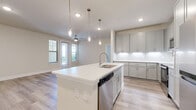 This well-appointed single-floor residence showcases a thoughtfully designed open-concept layout with contemporary finishes throughout. The kitchen serves as the centerpiece of the living space, featuring a generously sized island with an integrated sink, sleek white quartz countertops, soft-gray shaker-style cabinetry with brushed nickel hardware, a white tile backsplash with under-cabinet lighting, and a full suite of stainless steel Whirlpool appliances including a range, over-the-range microwave, dishwasher, and refrigerator. Elegant pendant lighting hangs above the island, adding a refined touch. The open living and dining area is bathed in natural light from multiple windows and a glass door that leads to an outdoor balcony, complemented by light wood-look flooring and a ceiling fan. The bedroom is carpeted for comfort, offers outdoor views through a large window, and includes access to a walk-in closet with wire shelving. The bathroom is finished with matching gray cabinetry, quartz countertops, a walk-in shower, and wood-look flooring. A dedicated laundry room houses a full-size top-load washer and dryer with overhead wire shelving for added storage. The overall ambiance is bright, clean, and modern.