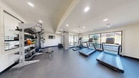 Located at 409 E William Cannon Dr in Austin, TX, this well-appointed fitness center offers a comprehensive workout experience within a single, efficiently designed exercise room. The space features a clean, modern aesthetic with crisp white walls, dark durable flooring, and recessed lighting that keeps the environment bright and energizing.

Large picture windows along multiple walls flood the room with natural light and provide views of the surrounding outdoor greenery, creating an open and motivating atmosphere. A stone-accented column adds an architectural touch that complements the contemporary design.

The fitness floor is thoughtfully organized into distinct training zones. A cardio section houses multiple treadmills and an elliptical machine positioned directly in front of the expansive windows. The strength training area features a multi-station cable machine system with TRX suspension trainers, a weight bench, and a well-stocked rack of dumbbells. A dedicated functional training zone includes medicine balls, kettlebells, slam balls, and plyo boxes for dynamic workouts.

A wall-mounted flat-screen television provides entertainment during workouts, while built-in ventilation ensures comfort throughout training sessions. The space is designed to accommodate a variety of fitness routines, making it a versatile and fully equipped facility for residents of the Austin community.