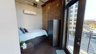 Located at 818 Georgia Ave in the heart of downtown Chattanooga, TN, this single-floor urban loft-style space masterfully blends historic character with modern functionality. The defining architectural feature is the richly textured exposed brick that lines the walls throughout, lending an authentic industrial aesthetic to the entire space.

The sleeping area is a compact yet well-appointed room featuring dark wood-toned furnishings, including a platform bed with built-in storage drawers and a tall wardrobe cabinet. A wall-mounted mini-split HVAC unit ensures year-round climate control, and a large glass door opens to a balcony with direct views of the surrounding downtown cityscape.

The living lounge area is a standout highlight of the space, anchored by two dramatic arched windows that flood the room with natural light and frame sweeping views of Chattanooga's downtown buildings and skyline. A teal sofa, patterned area rug, and casual floor cushions create a relaxed, inviting atmosphere. The exposed brick backdrop and dark hardwood flooring reinforce the loft's industrial-chic character.

The full bathroom is clean and contemporary in design, featuring dark cabinetry, a vanity with under-sink storage, a tiled walk-in shower, and a window that brings in a touch of natural light. The overall ambiance of this space is modern urban living rooted in historic architectural charm.