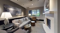 This single-floor space presents a refined, contemporary design centered around a well-appointed lounge area. The interior is finished in a neutral palette of warm grays and taupe tones, creating a calm and professional ambiance throughout. Recessed ceiling lighting and strategically placed floor and table lamps provide layered illumination, complementing the natural light that filters in through large gridded windows overlooking lush greenery outside.

The lounge area is furnished with a generous light gray sofa, coordinating armchairs in charcoal and brown upholstery, and round leather ottomans in a metallic bronze finish. Dark-framed side tables and a built-in low credenza along one wall add functional elegance. Abstract artwork is displayed prominently on the walls, contributing to the sophisticated atmosphere.

A fireplace serves as a focal point in one section of the space, framed by clean white millwork and accented by framed artwork above. The entryway features polished concrete flooring and dark wood-framed glass doors, transitioning smoothly into the carpeted lounge. The overall design balances comfort with a polished, upscale aesthetic suited to a professional or hospitality environment.