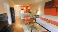 This single-floor residence showcases a thoughtfully designed open-concept layout that seamlessly blends the living, dining, and kitchen areas into one cohesive and welcoming space. Warm hardwood flooring flows throughout, anchoring the interior with a sense of continuity and warmth.

The kitchen is a standout feature, finished with rich cabinetry in a bold orange-toned wood, complemented by granite countertops, stainless steel appliances, and a built-in microwave. A peninsula with bar-height seating offers a casual dining option and serves as a natural divider between the kitchen and the main living area. Pendant lighting above the peninsula adds a refined decorative touch, while track lighting illuminates the kitchen workspace effectively.

The living area is open and airy, centered around a glass-topped coffee table and accented with a tufted cognac leather accent chair that adds a pop of color against the neutral walls. A clean white sofa, coordinating area rug, and a tasteful desk nook along one wall round out the space with functional elegance.

Near the entryway, a welcoming foyer with a white paneled door and a built-in shelving unit provides a practical and organized entry point. The overall ambiance is modern yet warm, with bold color accents and thoughtful furnishings creating a vibrant yet comfortable atmosphere.