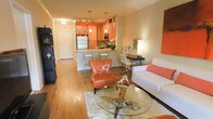 This single-floor residence showcases a thoughtfully designed open-concept layout that seamlessly blends the living, dining, and kitchen areas into one cohesive and welcoming space. Warm hardwood flooring flows throughout, anchoring the interior with a sense of continuity and warmth.

The kitchen is a standout feature, finished with rich cabinetry in a bold orange-toned wood, complemented by granite countertops, stainless steel appliances, and a built-in microwave. A peninsula with bar-height seating offers a casual dining option and serves as a natural divider between the kitchen and the main living area. Pendant lighting above the peninsula adds a refined decorative touch, while track lighting illuminates the kitchen workspace effectively.

The living area is open and airy, centered around a glass-topped coffee table and accented with a tufted cognac leather accent chair that adds a pop of color against the neutral walls. A clean white sofa, coordinating area rug, and a tasteful desk nook along one wall round out the space with functional elegance.

Near the entryway, a welcoming foyer with a white paneled door and a built-in shelving unit provides a practical and organized entry point. The overall ambiance is modern yet warm, with bold color accents and thoughtful furnishings creating a vibrant yet comfortable atmosphere.