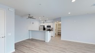 This single-floor space showcases a clean, contemporary design centered around an open-concept living and kitchen area. The kitchen is equipped with stainless steel appliances, including a refrigerator and microwave, and features crisp white cabinetry that extends to the upper walls, providing ample storage. A peninsula with a light-toned countertop separates the kitchen from the living area, offering additional prep space and a natural gathering point. Pendant lighting hangs above the peninsula, complementing the recessed ceiling lights that illuminate the space evenly throughout. Light hardwood-style flooring runs continuously across the open area, contributing to a cohesive and airy feel. A ceiling fan in the living area adds both functionality and a subtle modern touch. The walls are finished in a soft, neutral tone that enhances the brightness of the interior. Visible beyond the kitchen is a dedicated walk-in pantry or storage room, adding notable practical value to the layout. The overall ambiance is clean, well-lit, and functional, with a straightforward modern aesthetic suited to everyday living.