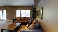 This single-floor space presents a thoughtfully designed living area that combines comfort and style through a cohesive, earthy color palette. The main living room is anchored by plush, dark leather and suede sofas adorned with decorative pillows in warm brown and chocolate tones, arranged around a rustic wooden coffee table to create an inviting seating arrangement. Recessed lighting in the ceiling provides consistent, ambient illumination throughout the room, complementing the natural light that filters in through a set of horizontal windows along one wall. The walls are finished in a rich, warm tan, lending the space a grounded and cozy atmosphere. A tall, lush indoor plant adds a natural, organic element to the corner of the room, contributing to the welcoming ambiance. A curated gallery of framed artwork lines one wall, adding visual interest and a personal touch to the décor. The textured accent wall further enhances the layered aesthetic of the space. Overall, this space exudes a relaxed yet refined character, well-suited for both quiet relaxation and casual social gatherings.