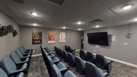 Located at 7856 S State Rt 48 in Maineville, Ohio, this single-floor private screening room delivers an authentic cinematic experience in a compact, purpose-built setting. The room is designed to mirror a traditional movie theater, with rows of blue upholstered theater seats arranged in a tiered, stadium-style layout, each equipped with built-in cup holders for added comfort. A large flat-panel display screen mounted at the front of the room serves as the focal point, providing a clear sightline from every seat in the house. The walls are painted in a deep, neutral gray that enhances the immersive atmosphere, while recessed ceiling lighting and a drop tile ceiling provide controlled, dimmable illumination ideal for screenings. Classic movie posters—including titles such as Traffic, Grease, and Forrest Gump—are framed and displayed along the walls, adding a nostalgic cinematic charm. Decorative film reel clusters are artfully arranged along the side and rear walls, reinforcing the theater theme throughout. Patterned carpet covers the floor, contributing to the room's acoustic and aesthetic qualities. A subwoofer is positioned near the seating area to support the audio setup. The space also features a popcorn machine near the entrance, completing the full movie-going experience.