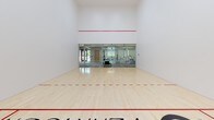 The Tennyson racquetball court is a purpose-built, single-level athletic facility designed to meet the demands of competitive and recreational play alike. The court features a pristine, light-toned hardwood floor with clearly marked red boundary and service lines, providing excellent traction and visual clarity during play. The surrounding walls are smooth, bright white, ensuring optimal ball visibility from all angles, while a bold red out-of-bounds line spans the full width of the front wall at regulation height.

One of the court's most striking architectural elements is its full-width glass back wall, composed of frameless glass panels that offer complete transparency into the adjacent fitness area beyond. This design not only provides a modern, open aesthetic but also allows spectators to observe play from outside the court. The fitness area visible through the glass is well-equipped with stationary bikes and other exercise equipment.

The ceiling soars to an impressive height, creating an expansive, airy atmosphere essential for unobstructed play. The clean, minimalist design of the space ensures there are no distracting elements, keeping the focus firmly on the game. The court floor is branded with "The Tennyson" lettering, reinforcing its identity as a premium amenity. Overall, this racquetball court combines functional design with a polished, contemporary aesthetic.