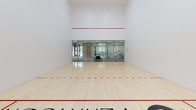 The Tennyson racquetball court is a purpose-built, single-level athletic facility designed to meet the demands of competitive and recreational play alike. The court features a pristine, light-toned hardwood floor with clearly marked red boundary and service lines, providing excellent traction and visual clarity during play. The surrounding walls are smooth, bright white, ensuring optimal ball visibility from all angles, while a bold red out-of-bounds line spans the full width of the front wall at regulation height.

One of the court's most striking architectural elements is its full-width glass back wall, composed of frameless glass panels that offer complete transparency into the adjacent fitness area beyond. This design not only provides a modern, open aesthetic but also allows spectators to observe play from outside the court. The fitness area visible through the glass is well-equipped with stationary bikes and other exercise equipment.

The ceiling soars to an impressive height, creating an expansive, airy atmosphere essential for unobstructed play. The clean, minimalist design of the space ensures there are no distracting elements, keeping the focus firmly on the game. The court floor is branded with "The Tennyson" lettering, reinforcing its identity as a premium amenity. Overall, this racquetball court combines functional design with a polished, contemporary aesthetic.
