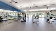 Located at 8601 Chatsfield Way in Hyattsville, MD, this fitness center offers a generously sized, single-floor space designed to support a comprehensive workout experience. The open floor plan provides ample room for members to move freely between equipment zones without feeling crowded.

The interior features a bold and energizing color scheme, with vivid yellow and blue accent walls complemented by crisp white ceilings. Large wall-to-wall mirrors line one side of the facility, visually expanding the space and allowing users to monitor their form during exercise. Ceiling fans are mounted throughout, promoting air circulation and comfort during intense workouts.

The cardio zone is well-stocked with treadmills, stair climbers, and stationary bikes, many equipped with individual screens for entertainment or tracking. The strength training area includes cable machines, a Smith machine, weight benches, free weights, battle ropes, and stability balls, providing a thorough range of resistance training options. Flat-screen TVs are mounted on the walls for additional viewing convenience.

Natural light pours in through large windows and glass doors that line the perimeter, offering pleasant views of the surrounding greenery. The durable rubber flooring throughout is well-suited for heavy equipment and high-impact activities. Overall, the facility delivers a bright, functional, and motivating environment for fitness enthusiasts of all levels.