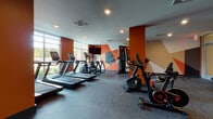 Located at 67 Old Amboy Rd in Old Bridge, NJ, this fitness center is designed to provide a comprehensive workout environment within a single-floor layout. The space is defined by its bold aesthetic, featuring warm terracotta-orange walls accented by a striking geometric mural with angular shapes in orange, tan, and grey tones, creating an energetic and motivating atmosphere throughout.

The cardio zone is positioned along a wall of large windows, allowing natural light to fill the area while offering outdoor views. It is equipped with multiple treadmills and elliptical trainers, each featuring integrated digital displays. Matrix-brand stationary bikes and a recumbent cycle provide additional low-impact cardio options.

The strength training area is well-appointed with a multi-station cable machine, a full rack of Matrix dumbbells, adjustable benches, and a wall-mounted mirror that enhances the sense of space. Additional equipment including stability balls and resistance tools is neatly organized on dedicated storage racks along the perimeter.

Recessed ceiling lighting throughout ensures consistent illumination, while durable rubber flooring provides safety and comfort underfoot. A mounted flat-screen television and posted fitness center rules complete this practical, purpose-built facility.