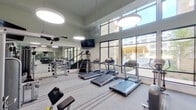 This fitness center is a thoughtfully designed workout facility that combines functionality with an open, airy aesthetic. The space boasts soaring double-height ceilings that create an expansive atmosphere, complemented by large floor-to-ceiling windows along one wall that flood the interior with natural light and offer views of an outdoor courtyard and pool area beyond.

The cardio zone features multiple treadmills, elliptical cross-trainers, and a stair climber, all positioned to take advantage of the abundant natural light and outdoor views. Oversized pendant light fixtures hang from the high ceiling, providing additional illumination throughout the space.

A dedicated strength training area offers a comprehensive selection of equipment, including a cable machine, a leg extension/curl machine, a weight bench with a rack, and a fully stocked dumbbell rack with a wide range of weights. Full-length mirrors line one wall, visually expanding the space and allowing users to monitor their form during workouts. Multiple wall-mounted flat-screen televisions are positioned throughout the facility for entertainment.

The flooring features a durable striped carpet, and the neutral color palette of soft white and gray tones contributes to a clean, modern ambiance. Black-framed glass double doors serve as the main entry point, reinforcing the contemporary design aesthetic of this well-appointed fitness facility.