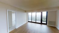 This single-floor unit features an open and airy living area defined by warm hardwood-style flooring that runs throughout the main space. The most striking architectural element is the expansive wall of floor-to-ceiling sliding glass doors, which flood the interior with abundant natural light and provide direct access to a private balcony. The balcony is framed by a classic dark metal railing and overlooks a lush, tree-filled landscape, creating a serene and natural backdrop. The interior walls are finished in a neutral, off-white palette that complements the light tones of the flooring, contributing to a clean and versatile aesthetic. A textured ceiling adds subtle visual character to the space. An adjacent doorway visible from the main living area suggests additional rooms beyond, offering separation between living and other functional spaces. The open layout of the living area allows for flexible furniture arrangement, making it adaptable to a variety of lifestyle needs. Overall, the unit balances simplicity with functionality, highlighted by its generous natural light and pleasant outdoor views.