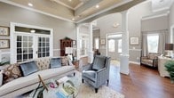 This sophisticated single-floor space presents a refined blend of classic architectural details and upscale interior design. Rich hardwood flooring flows throughout, anchoring each area with warmth and continuity. The main gathering area commands attention with its impressively high ceilings, accented by coffered beam detailing and recessed lighting that illuminate the space evenly. Tall, grid-paned French doors and large arched windows line the walls, drawing in generous natural light and offering views to the outdoors.

Stately white columns with square pedestals define zones within the open layout, adding a sense of structure without sacrificing the airy, open feel. Neutral gray walls paired with crisp white crown molding and chair-rail wainscoting lend an air of timeless elegance. The seating area is appointed with a tufted linen sofa, quilted velvet accent chairs, and a glass-top coffee table arranged atop a soft, patterned area rug.

Additional areas feature an ornate iron console table, wall-mounted display screens, and curated decorative accents including ceramic urns and framed artwork. Multiple doorways with frosted glass panels lead to adjacent private offices or ancillary rooms. The overall ambiance is polished and welcoming, reflecting a professional yet comfortable environment suited for community gatherings or reception purposes.