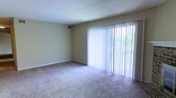 This single-floor space features a straightforward, functional layout with a focus on simplicity and practicality. The main living area is finished with wall-to-wall carpeting in a neutral beige tone, providing a soft underfoot feel that complements the light-colored walls throughout. A standout feature of the room is the large sliding glass door, which spans a significant portion of one wall and is dressed with full-length vertical blinds. This door serves as the primary source of natural light, allowing an abundance of daylight to filter into the space while offering views of the greenery visible outside. The sliding door also provides direct access to an outdoor area, enhancing the sense of connection to the exterior. The walls are painted in a warm, neutral beige tone that contributes to an open and airy feel. A ceiling-mounted HVAC vent is visible, indicating a central air system for climate control. The clean, uncluttered layout of the space makes it highly adaptable to various furnishing configurations, offering flexibility for a range of uses. White baseboards provide a clean finish throughout the room.
