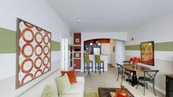 This single-floor residence offers a thoughtfully designed open-concept layout that seamlessly connects the living, dining, and kitchen areas into one inviting and functional space. The kitchen is a standout feature, highlighted by a arched entryway with warm terracotta and deep red accent walls, dark cabinetry, stainless steel appliances, and a raised breakfast bar with sleek green bar stools — perfect for casual dining or entertaining. Adjacent to the kitchen, a dedicated dining area accommodates a round table with modern cross-back chairs, creating an intimate setting for meals. The living area is bright and welcoming, featuring a white sofa accented with green and orange throw pillows, contributing to the space's warm, earthy color palette. The walls are adorned with bold horizontal green and white stripes, complemented by eye-catching artwork, adding a playful yet polished character throughout. Light-toned flooring and white ceilings enhance the sense of openness, while the flowing layout ensures easy movement between spaces. A hallway visible in the background suggests additional private rooms, completing this well-appointed and colorfully expressive home.