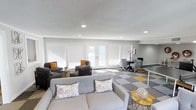 This single-floor space offers a well-organized, open-concept layout that combines comfortable lounging with functional workspace areas. The interior is finished with neutral grey walls and a textured white ceiling with recessed lighting, creating a clean and well-lit environment throughout.

The lounge area features a grey sofa adorned with floral and patterned throw pillows, complemented by two dark upholstered armchairs with orange accent cushions. A sleek glass-and-gold side table adds a contemporary touch to this relaxed seating zone.

Adjacent to the lounge, a dedicated workspace area is equipped with office chairs and a desk, making the space suitable for both collaborative and focused work. The flooring is a distinctive modular carpet in a checkered pattern of grey, white, and yellow tones, adding visual interest and helping to define each functional zone.

Large windows with white horizontal blinds and a central set of French doors allow abundant natural light to fill the room while providing access to the exterior. The overall ambiance is modern and inviting, balancing productivity with comfort.