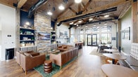 Located at 1700 64th Avenue in Kenosha, Wisconsin, this single-floor clubhouse space masterfully blends rustic charm with modern design. The architecture is defined by dramatic vaulted ceilings with exposed wood planking and heavy timber trusses, complemented by bold black steel beam accents and visible ductwork that add an industrial edge. Wide-plank hardwood flooring flows throughout, unifying the open-concept interior.

The main lounge area anchors the space with a generous seating arrangement of rich cognac leather sofas and armchairs surrounding a round wood coffee table, all set against a striking floor-to-ceiling stacked stone feature wall with built-in shelving and a wall-mounted flat screen television. Abundant natural light pours in through an impressive two-story glass wall and French doors at the rear, offering views of the surrounding outdoors.

A well-appointed kitchen island with a light quartz countertop and leather swivel bar seating serves as a social hub, backed by a full service counter with cabinetry, a subway tile backsplash, stainless steel appliances, and open shelving. A separate dining area with sage green walls, wood beam columns, and a farmhouse-style table provides a more intimate setting. The overall ambiance is warm, inviting, and thoughtfully designed for community gathering.