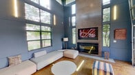 This contemporary amenity lounge is defined by its dramatic double-height ceilings and expansive floor-to-ceiling windows that flood the interior with natural light and offer lush greenery views. The main lounge area features sleek, low-profile modular white sectional sofas arranged around a circular white coffee table, all set atop polished concrete flooring that adds an industrial-chic character to the space. A towering concrete accent wall serves as the room's focal point, housing a wall-mounted flat-screen television above a modern linear fireplace, creating a bold and inviting atmosphere. Vertical linear light fixtures are mounted along the blue-grey paneled walls, adding a warm, contemporary glow throughout. The lounge opens directly to an outdoor covered patio through large industrial-style overhead doors that retract fully, blurring the boundary between indoors and outdoors. The exterior area features a stone-clad outdoor kitchen equipped with stainless steel grills and a covered pergola, with views extending to a pool area beyond. The combination of high-design interior finishes, abundant natural light, and effortless indoor-outdoor connectivity makes this space a highly functional and visually striking communal gathering area.