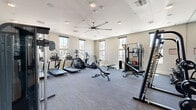 Located in Surprise, Arizona, this fitness center offers a comprehensive workout environment designed to support a wide range of exercise routines. The space is thoughtfully laid out across a single floor, providing ample room to move freely between equipment zones without feeling crowded.

The cardio section is well-stocked with multiple treadmills, elliptical machines, a stair climber, and a stationary bike, each equipped with individual screens for an engaging workout experience. A dedicated strength training area includes a multi-functional cable machine, adjustable weight bench, and a full dumbbell rack, catering to both beginners and experienced athletes.

The room is finished with neutral gray tones and plush carpet flooring that absorbs sound and adds comfort underfoot. A large ceiling fan anchors the space visually while promoting consistent air circulation throughout. Full-wall mirrors on one side enhance the sense of openness and allow users to monitor their form during exercises.

Natural light pours in through multiple large windows positioned along the walls, offering views of the surrounding community and keeping the atmosphere bright and energizing. Recessed lighting supplements the natural light for evening use. A wall-mounted television adds an additional entertainment option. The overall ambiance is clean, modern, and purposefully designed for effective fitness use.