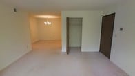 This single-floor space is characterized by its straightforward, functional layout and neutral design palette. The main area is finished with wall-to-wall light beige carpeting that extends throughout, lending a cohesive and understated feel to the interior. The walls are painted in a clean, off-white tone that reflects light evenly across the space.

A prominent built-in closet is centrally visible, featuring a bi-fold door and an interior hanging rod, offering practical storage. Adjacent to this closet, a secondary door provides access to another area of the unit. The closet's open design reveals a generous depth, suitable for a variety of storage needs.

An adjoining room is visible through an open passageway, distinguished by a chandelier-style light fixture that adds a modest decorative touch to an otherwise simply appointed space. The chandelier features multiple arms and provides warm overhead lighting to that area.

Overall, the space presents a clean, move-in ready environment with consistent flooring, neutral tones, and adequate storage, suited to a variety of furnishing arrangements.