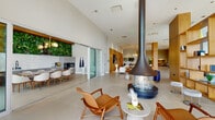 Located at 900 Pacific St in Stamford, Connecticut, this sophisticated amenity floor offers a rich array of curated spaces designed for relaxation, wellness, and socializing. The expansive main lounge is defined by soaring ceilings, large-format tile flooring, and dramatic floor-to-ceiling windows that flood the interior with natural light and frame views of the surrounding outdoor terrace. A striking suspended fireplace serves as the centerpiece of the lounge, complemented by mid-century modern furnishings, warm wood accents, and elegant pendant lighting. An adjoining dining and kitchen area features a vibrant green living wall, adding a natural, organic touch to the contemporary design. A game and reading nook offers a round table beneath a sculptural chandelier, flanked by a built-in wood shelving display. The spa area is a standout destination, highlighted by a serene indoor water feature with a lush planted surround, a glass-enclosed sauna with teak wood finishes, a dedicated relaxation lounge, and a full-service salon with glass-partitioned interiors. A gold-railed staircase connects the levels while overlooking the tranquil water installation. Throughout, the design balances refined materials with an open, welcoming ambiance.
