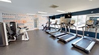 This fitness center is designed to support a full range of workout routines within a spacious, single-floor layout. The space is divided into distinct functional zones, seamlessly connected to create an open and energizing environment. The cardio area is lined with a row of TRUE treadmills, upright bikes, and elliptical machines positioned along large windows that allow abundant natural light and outdoor views, contributing to an invigorating workout atmosphere. Wall-mounted flat-screen TVs are positioned above the cardio equipment for added convenience. The strength training section features a variety of cable machines, weight stations, and benches suitable for both upper and lower body workouts. Free weights and additional resistance equipment are also available, offering flexibility for diverse training needs. The flooring throughout is durable rubber matting, ideal for high-traffic fitness use. Motivational typography murals in bold orange and navy tones cover feature walls, reinforcing an energetic and focused ambiance. Large mirrors extend along key wall sections, enhancing the sense of space. Recessed and flush-panel lighting provides consistent illumination throughout. French doors at one end open to an outdoor area, adding an element of connectivity to the exterior. The overall design is clean, modern, and purposefully laid out to maximize usability and motivation.