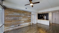 This single-floor space showcases a thoughtfully designed interior that blends rustic and contemporary elements seamlessly. The most commanding feature of the main living area is a full-length reclaimed wood accent wall, composed of varied plank tones ranging from warm honey to weathered grey, lending the space a distinctive character and visual depth. Underfoot, rich hardwood-style flooring runs throughout, complementing the natural textures of the accent wall and tying the design together cohesively.

A ceiling fan with sleek dark-toned blades is centrally mounted, providing both airflow and a modern finishing touch to the open living area. The layout flows effortlessly into an open-concept kitchen, which features dark cabinetry, pendant lighting in an industrial style, a stainless steel refrigerator, and a countertop bar with a sink — ideal for both everyday use and casual entertaining. Recessed lighting in the kitchen adds a warm, functional glow.

White-trimmed doorways and crisp white walls provide clean contrast against the darker design elements, maintaining a bright and airy atmosphere throughout. The overall ambiance balances modern minimalism with warm, textural details, creating a space that feels both stylish and inviting.