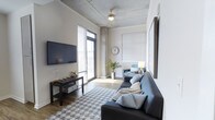 Located at 28 Woolfe St in Charleston, SC, this single-floor unit offers a compelling blend of industrial aesthetics and modern comfort in one of the city's most walkable and sought-after neighborhoods. The living area immediately draws attention with its exposed concrete ceiling, which lends a loft-like, urban character to the space. A ceiling fan adds both function and style beneath the raw overhead finish. Large windows fitted with horizontal blinds invite an abundance of natural light throughout the day, brightening the hardwood floors and neutral wall tones. The living room is thoughtfully laid out, accommodating a full sofa, accent seating, and a media console with a wall-mounted flat-screen television, all anchored by a geometric-patterned area rug. Touches of greenery and warm accent lighting contribute to a balanced, inviting ambiance. The overall design palette—cool grays, soft whites, and warm wood tones—creates a cohesive and contemporary aesthetic throughout. The space reflects the character of Charleston's historic Cannonborough-Elliotborough district, offering proximity to local dining, shopping, and cultural attractions while maintaining a stylish and functional interior environment.