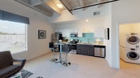 This contemporary urban loft showcases an industrial-chic aesthetic defined by striking exposed concrete beam ceilings that run throughout the space, lending it a raw yet refined character. The open-concept layout seamlessly connects the kitchen and living areas, creating a spacious, airy atmosphere. The kitchen is outfitted with stainless steel appliances, dark wood cabinetry, crisp white upper cabinets, and a soft teal tile backsplash that adds a pop of color. A bistro-style bar table with chrome stools provides a casual dining option within the kitchen space. The living area is bathed in natural light streaming through large windows dressed with blue valances and horizontal blinds, offering views of the surrounding greenery. High ceilings amplify the sense of openness, while a large decorative mirror enhances the brightness of the room. The bedroom features the same soaring exposed concrete ceilings and is complemented by a walk-in closet with built-in shelving. The bathroom is clean and modern, with a dark wood vanity, a single undermount sink, a large mirror, and light tile flooring, accented by teal décor for a cohesive, spa-like feel. Throughout the space, the design strikes a balance between industrial architecture and warm, contemporary furnishings.