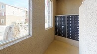 This mail area is a single-level communal space designed to serve the practical needs of a multi-unit residential community. The room is defined by its textured stucco walls finished in a warm tan tone, which are consistent throughout and lend the space a cohesive, straightforward aesthetic typical of residential common areas.

The central feature of the space is the extensive arrangement of individual metal mailboxes, organized across two large banks mounted directly into the stucco walls. The lockboxes are dark-finished and clearly numbered, providing secure mail access for a significant number of residents. The layout is efficient, maximizing wall space to accommodate the volume of units served.

A cork bulletin board is mounted on one wall, currently displaying a variety of community notices and informational flyers, serving as a communication hub for residents. The concrete flooring is durable and practical, suited to the functional nature of the space.

Natural light enters through multiple windows positioned along the walls, offering views of the surrounding residential exterior, including neighboring buildings. This lighting reduces the reliance on artificial illumination and adds a degree of openness to what is otherwise a compact, utilitarian space. The overall ambiance is clean, organized, and functional.