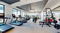Located at Waters Edge Village in Charlotte, NC, this expansive two-floor fitness center offers a comprehensive workout experience across generously sized levels. The main floor is anchored by a large cardio and strength training area outfitted with Matrix treadmills, ellipticals, stair climbers, and multi-functional cable machines. Full-length mirrors line the walls, enhancing the sense of space, while ceiling fans and recessed lighting ensure a comfortable environment. Large floor-to-ceiling windows flood the space with natural light and frame striking views of the surrounding waterfront and community grounds. A dedicated studio area on this level features a ballet barre, exercise balls, wall-mounted TV, and open floor space ideal for group fitness, yoga, or stretching. The upper floor continues the fitness theme with additional cardio equipment including rowing machines, spin bikes, and a functional training station, complemented by a wall-mounted TV and ample open space. A dramatic spiral staircase connects the two floors, while a striking arched window on the upper landing offers a picturesque view of the lake. The entry area features a teal built-in credenza and a tray ceiling detail, providing a polished welcome to the facility. Throughout, the design balances modern aesthetics with practical functionality.