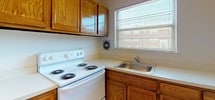 This single-floor unit located on Brynwood Gardens in Old Bridge, NJ, presents a straightforward and functional layout spread across several distinct areas. Throughout the living spaces and bedrooms, warm-toned hardwood flooring runs consistently, reflecting natural light that enters through multiple windows dressed with horizontal blinds. The white-painted walls and clean ceilings contribute to a bright, open atmosphere in each room.

The living area is generously proportioned, featuring baseboard heating and a wall-mounted air conditioning unit, ensuring year-round comfort. Natural light filters in through a wide multi-pane window, illuminating the polished hardwood floors. An additional room, suitable as a bedroom, mirrors this design with its own window and baseboard heating.

The kitchen is equipped with warm oak cabinetry both above and below the countertops, a white electric coil-top range, and a stainless steel sink beneath a window. A wall ventilation fan provides practical airflow in the cooking area.

The bathroom presents a distinctive retro character, fully tiled in light blue from floor to mid-wall, complemented by a matching blue tub and toilet. A wood-trimmed vanity with a white basin and a mirrored medicine cabinet complete the space. The textured ceiling and mosaic tile flooring add to the room's classic aesthetic.